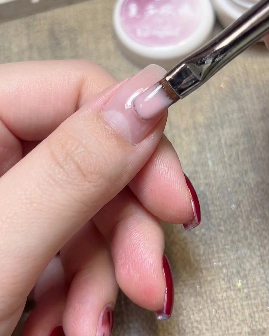 Gros plan sur un pinceau gel plat appliquant un vernis rose clair translucide sur un ongle naturel.