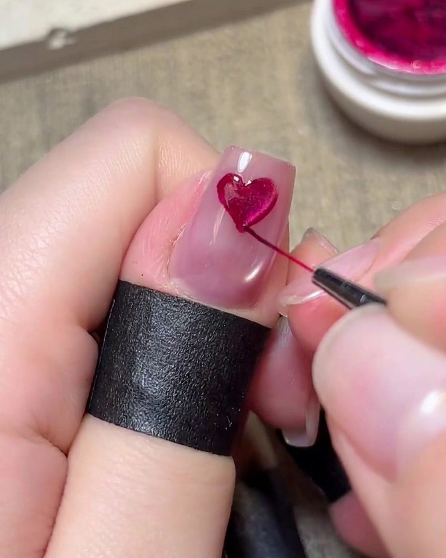 Un pinceau fin détail peignant soigneusement un cœur magenta sur un ongle rose clair.