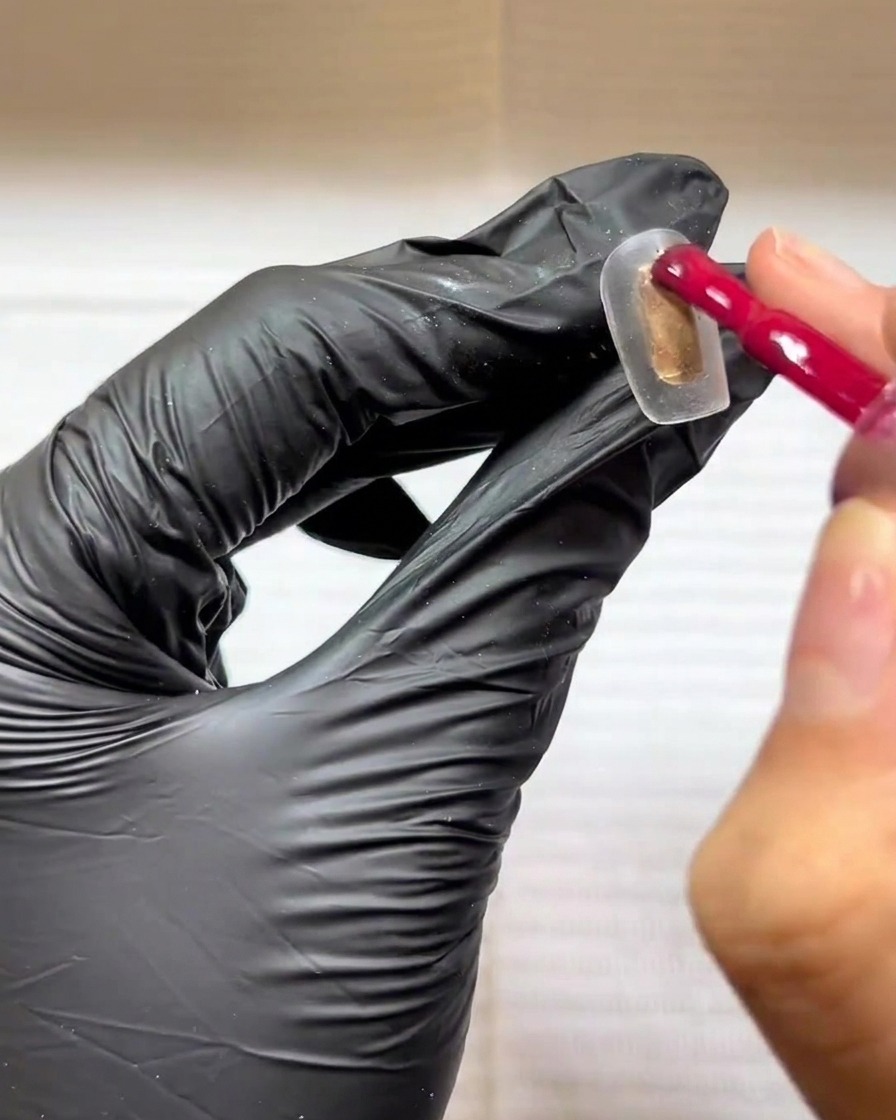Une main gantée utilisant un pinceau gel pour appliquer une couleur rouge rose sur une capsule transparente.