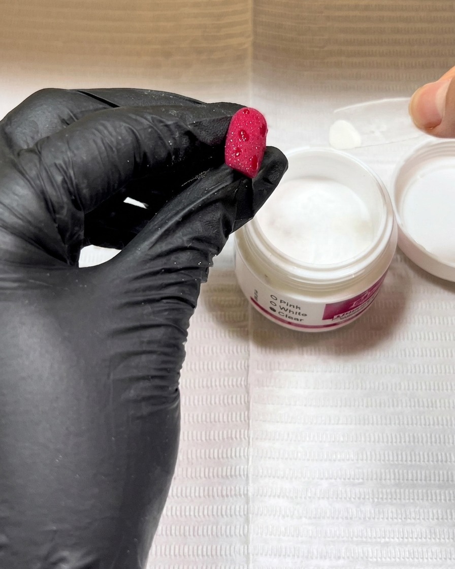 Mains gantées tenant un ongle rouge au-dessus dun pot de poudre transparente, saupoudrant la poudre sur des points de gel humide.
