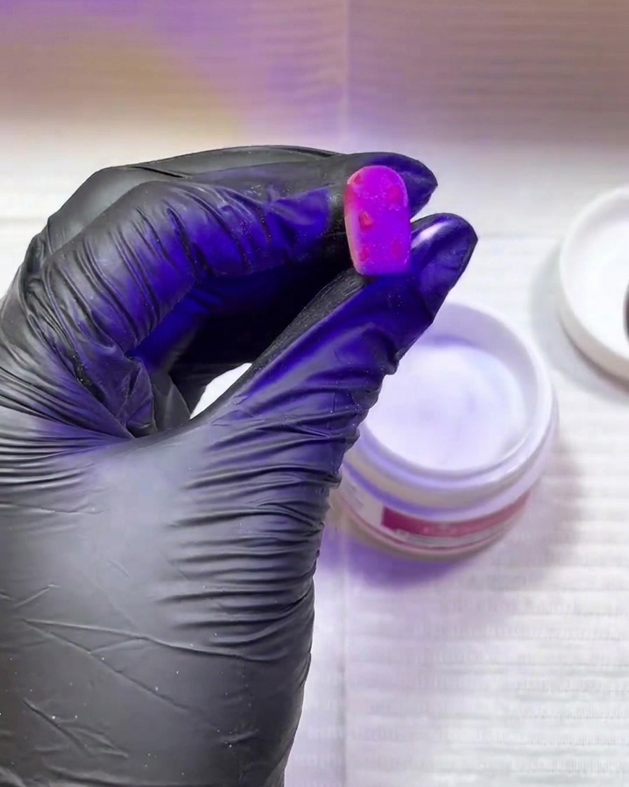 Une main portant un gant noir tenant un ongle rose vif avec des taches texturées sous une lampe de catalyse UV/LED violette.