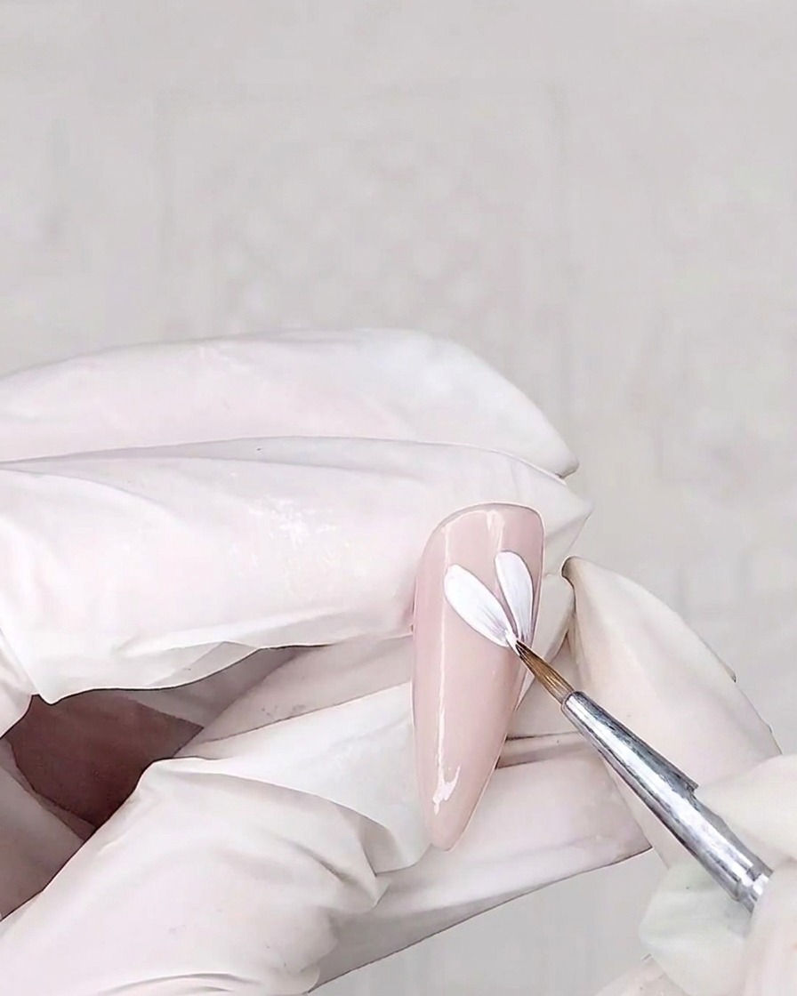 Un pinceau de détail peignant des pétales blancs sur un ongle enduit de rose.