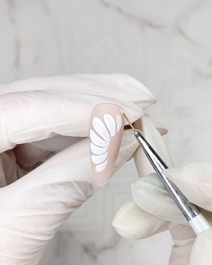 Gros plan dun pinceau liner appliquant une fine ligne de gel adhésif autour du bord dun pétale de marguerite blanc sur une base nude.