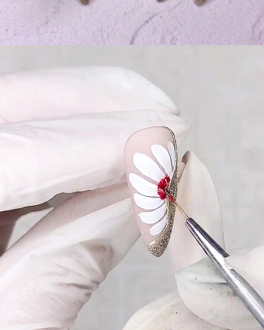 Un pinceau de détail fin appliquant du gel rouge au centre dun design de marguerite blanche sur un ongle.