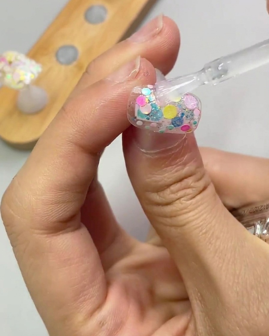 Un pinceau applique une couche transparente de gel de renforcement sur un ongle décoré de paillettes.