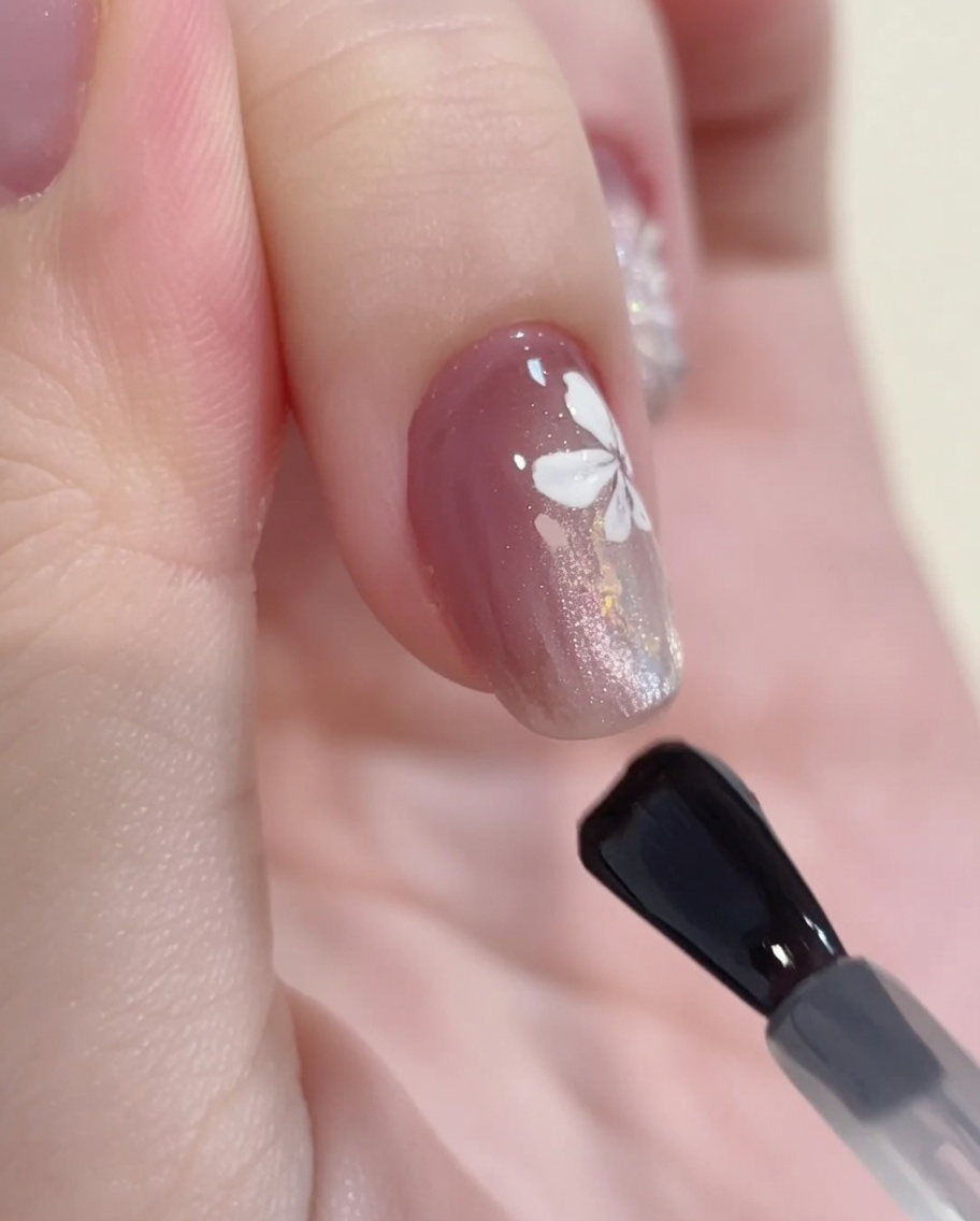 Un pinceau gel appliquant un top coat brillant sur un design final combinant cat-eye, paillettes et art floral.