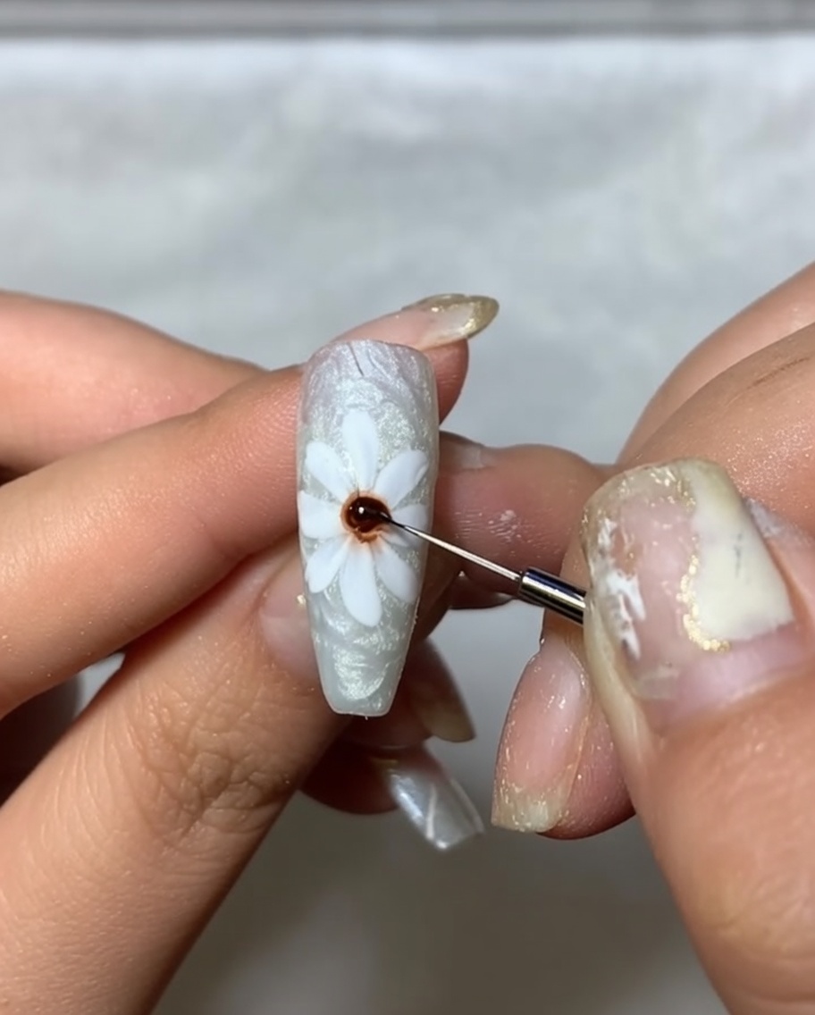 Gros plan dun ongle avec des pétales blancs peints, utilisant un pinceau fin pour appliquer un point de gel marron foncé au centre.