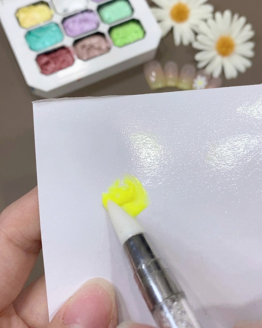 Un pinceau mélangeant le gel jaune et la base coat sur une surface blanche pour créer un mélange fin et homogène.