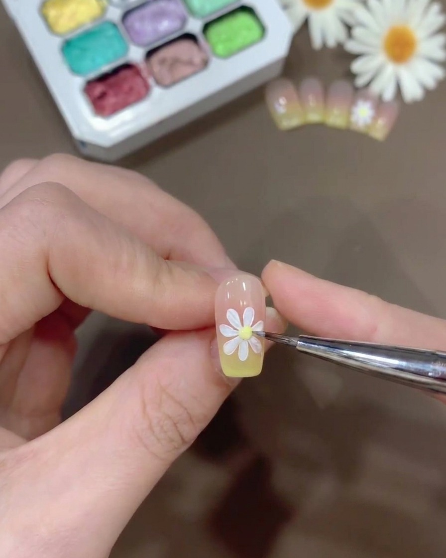 Une minuscule perle de gel jaune étant placée au centre dune marguerite blanche peinte sur un ongle.