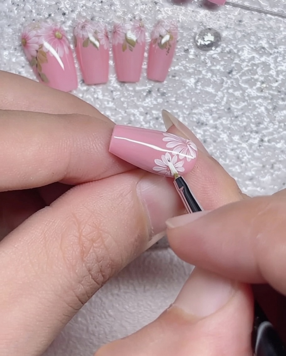 Un pinceau liner traçant soigneusement le périmètre des pétales de marguerite blancs sur un ongle rose.