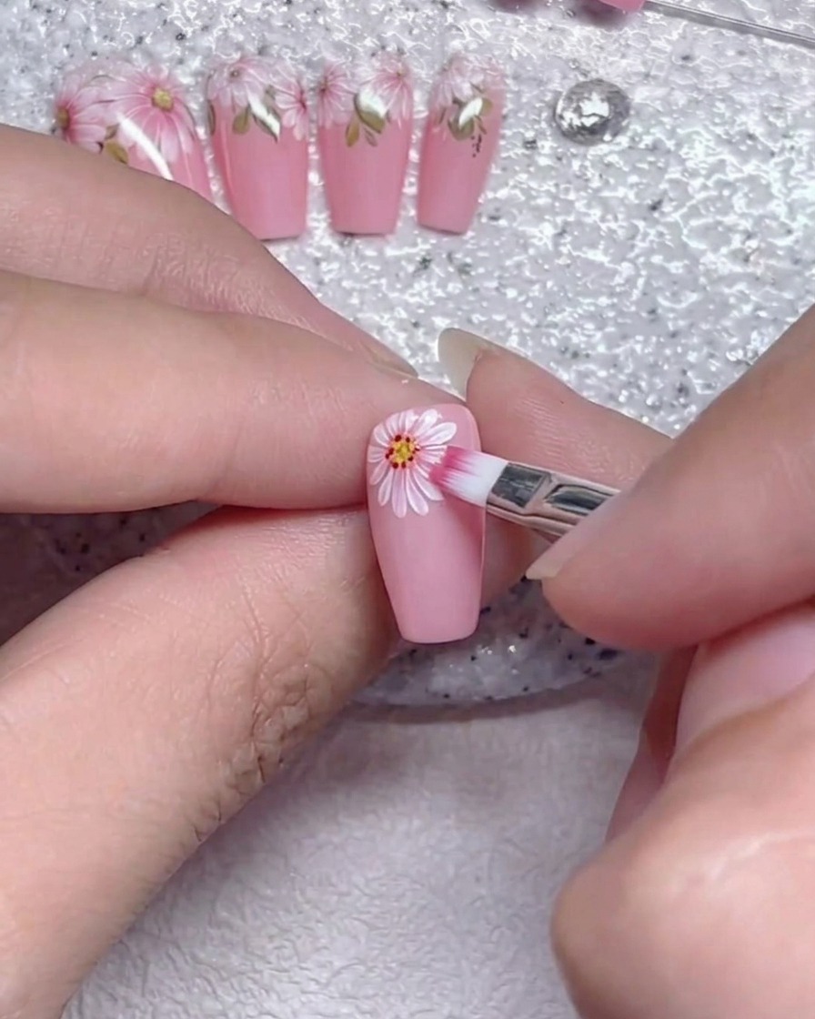 Un pinceau plat estompant doucement les bords des pétales de fleurs blancs sur un ongle rose.