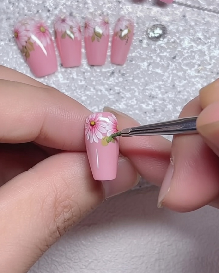 Gros plan dun pinceau liner ajoutant des détails de feuilles vertes à la base dune marguerite sur un ongle rose.