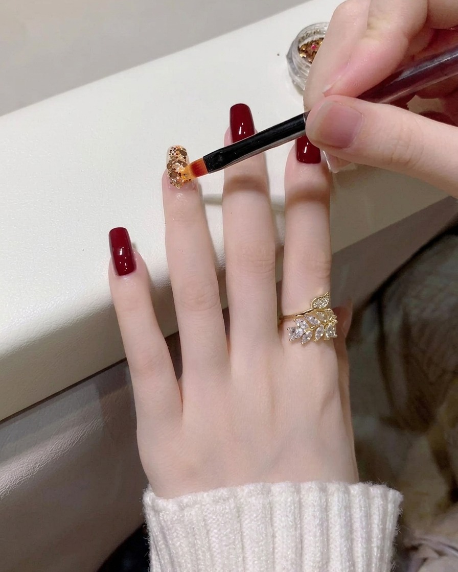 Gros plan sur un pinceau de précision déposant de petites paillettes dorées et holographiques sur un ongle recouvert dune base transparente.