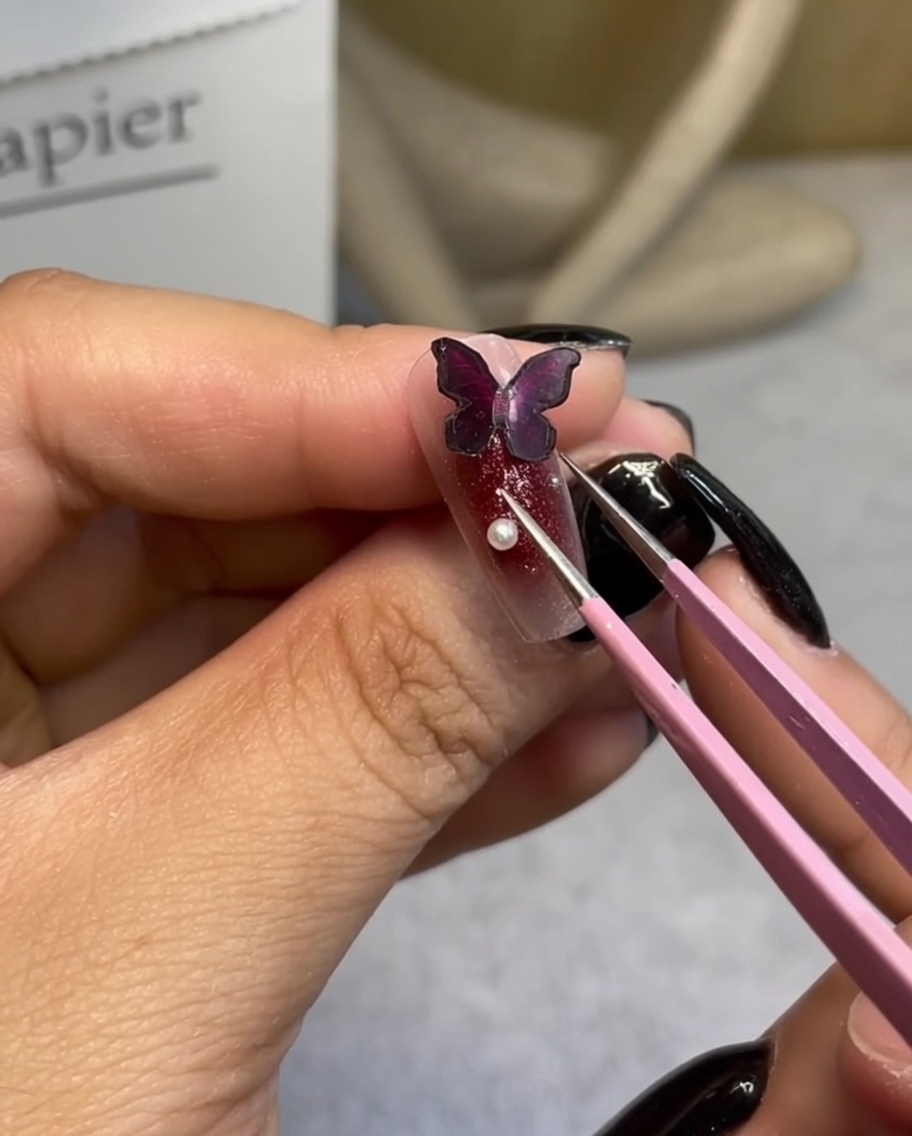 Une pince de précision plaçant une petite perle blanche près dun papillon.