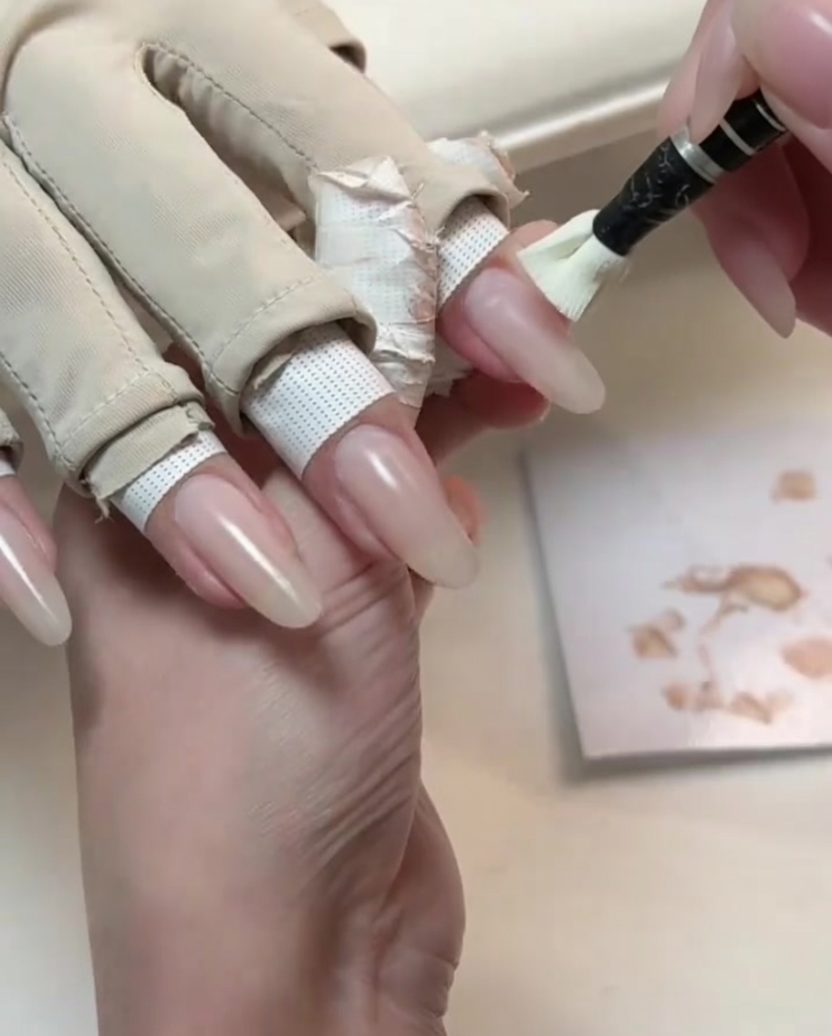 Un petit pinceau appliquant un vernis gel nude sur un ongle long de forme naturelle.