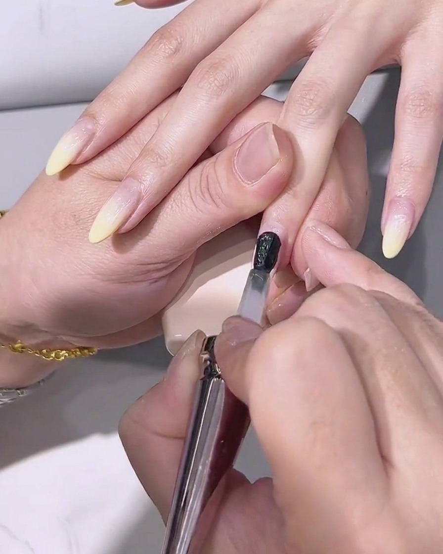 Un pinceau appliquant un top coat transparent et brillant sur longle au dégradé jaune citron.