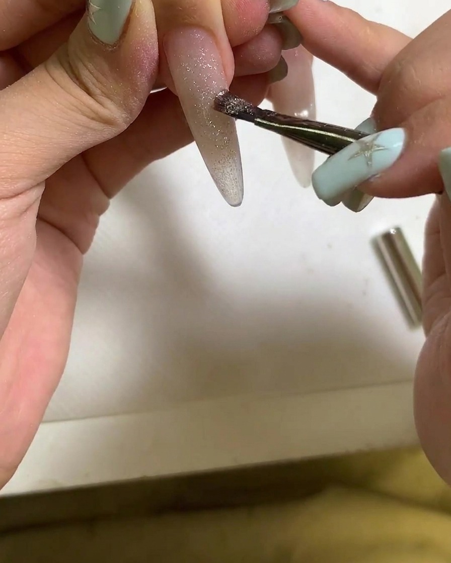 Un pinceau appliquant du vernis gel cat eye sur un ongle allongé.