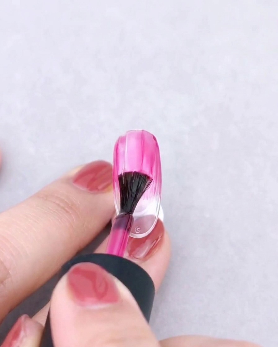 Un pinceau éventail estompant délicatement une couleur de gel rose vers le bas sur un ongle transparent pour créer un effet dégradé.