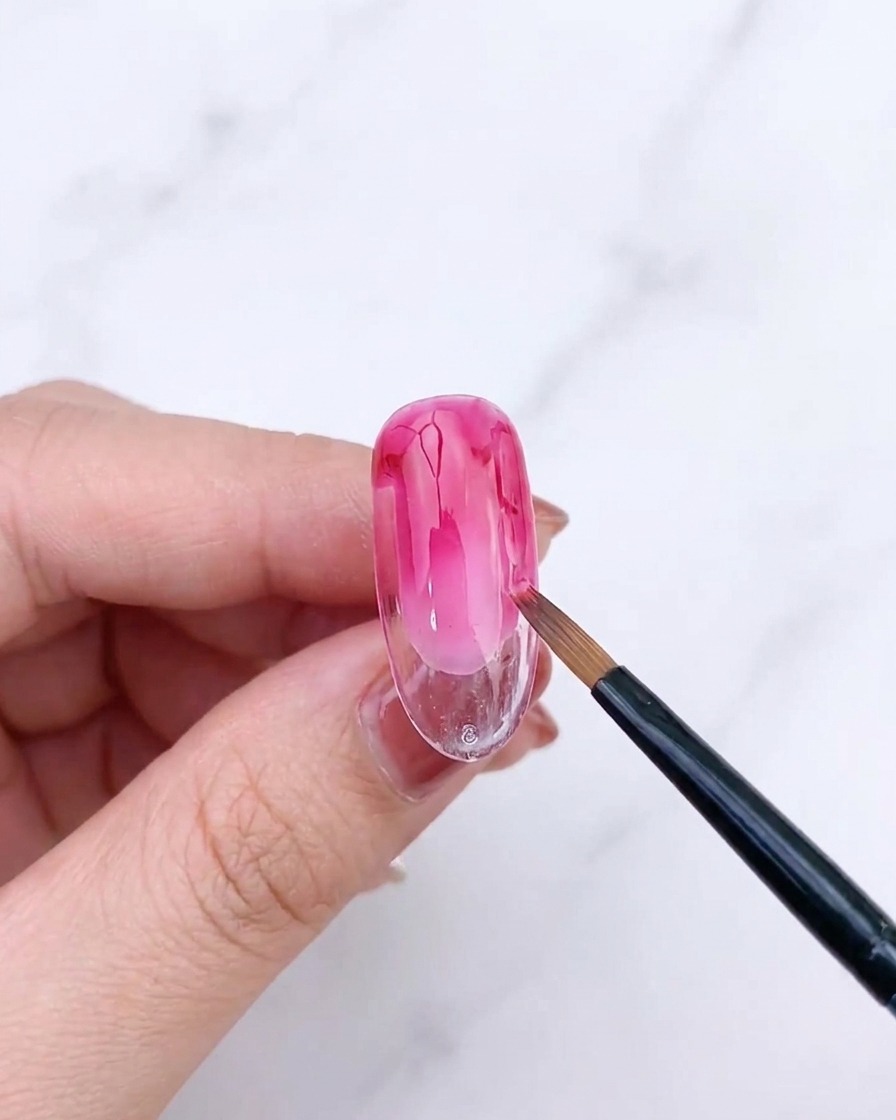 Un pinceau éventail estompant la seconde couche de gel rose sur un ongle pour affiner le dégradé.