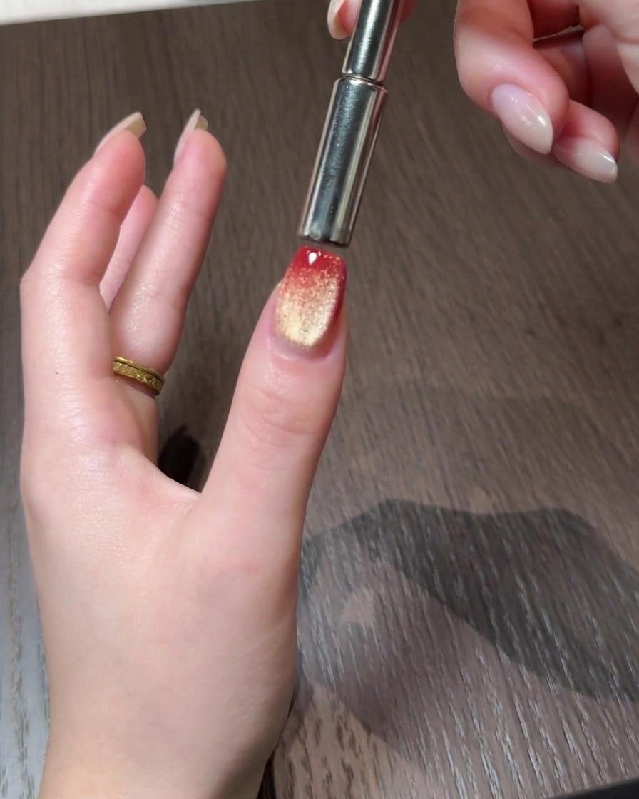 Un outil magnétique poussant les particules cat eye vers le bord arrière dun ongle en dégradé rouge et or.