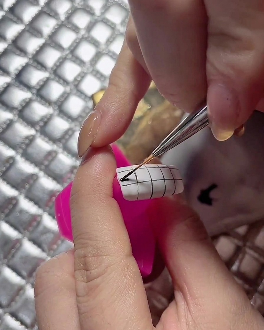 Un pinceau de détail colorant proprement les carrés alternés sur un ongle au motif quadrillé.