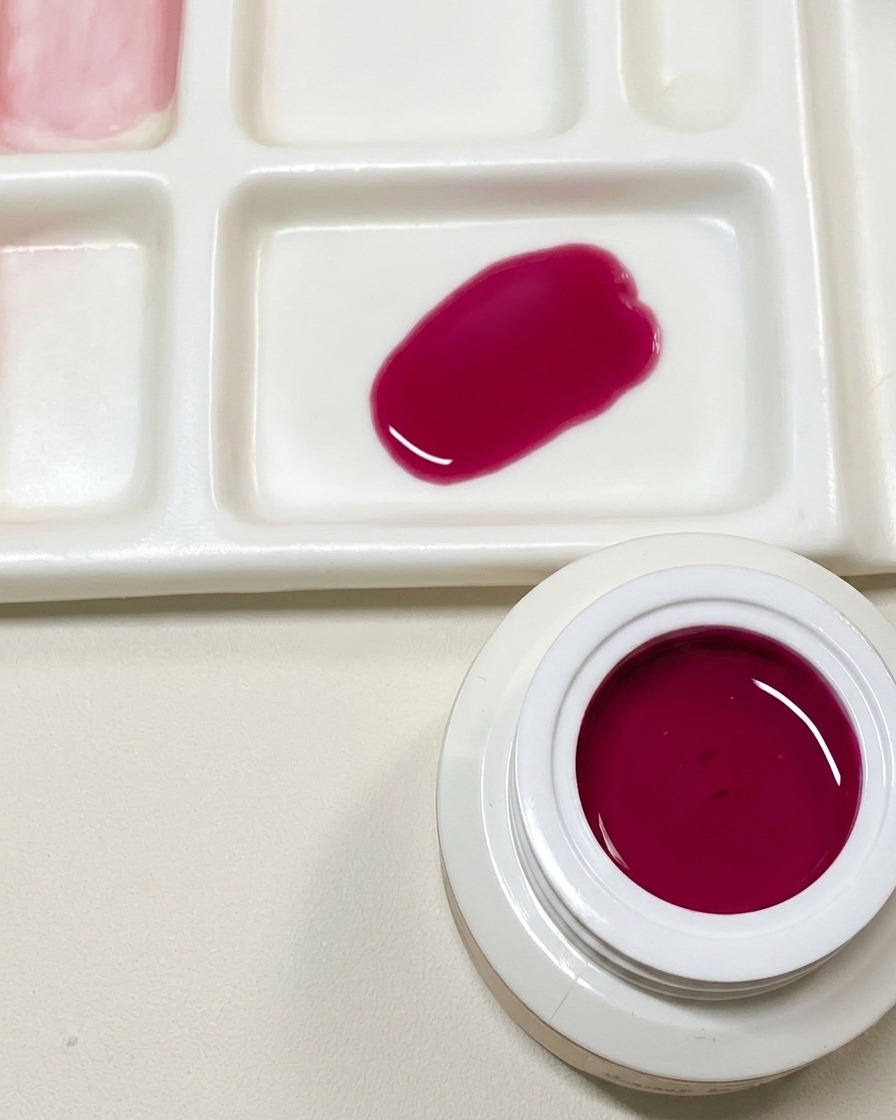 Une palette de mélange montrant un échantillon de vernis gel rose foncé personnalisé à côté dun pot ouvert de gel rouge fruit du dragon.