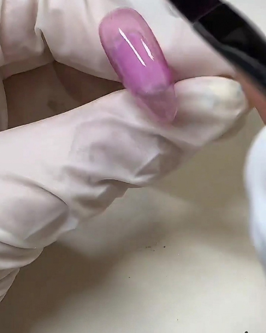 Une main gantée utilisant un pinceau à gel pour appliquer un vernis gel rose semi-transparent sur une capsule dongle transparente.