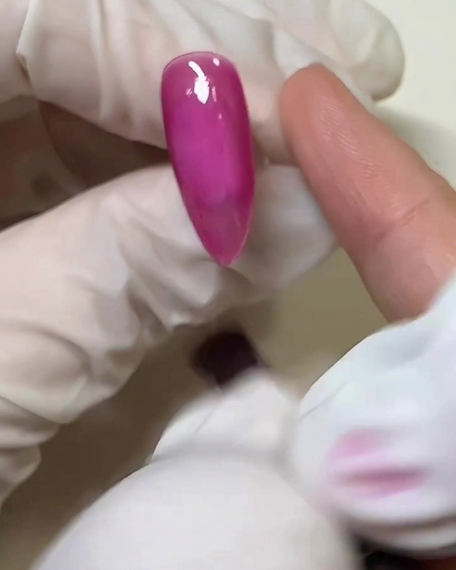 Gros plan dun doigt ganté appliquant une fine couche de colle solide transparente sur la surface rose de longle catalysé.