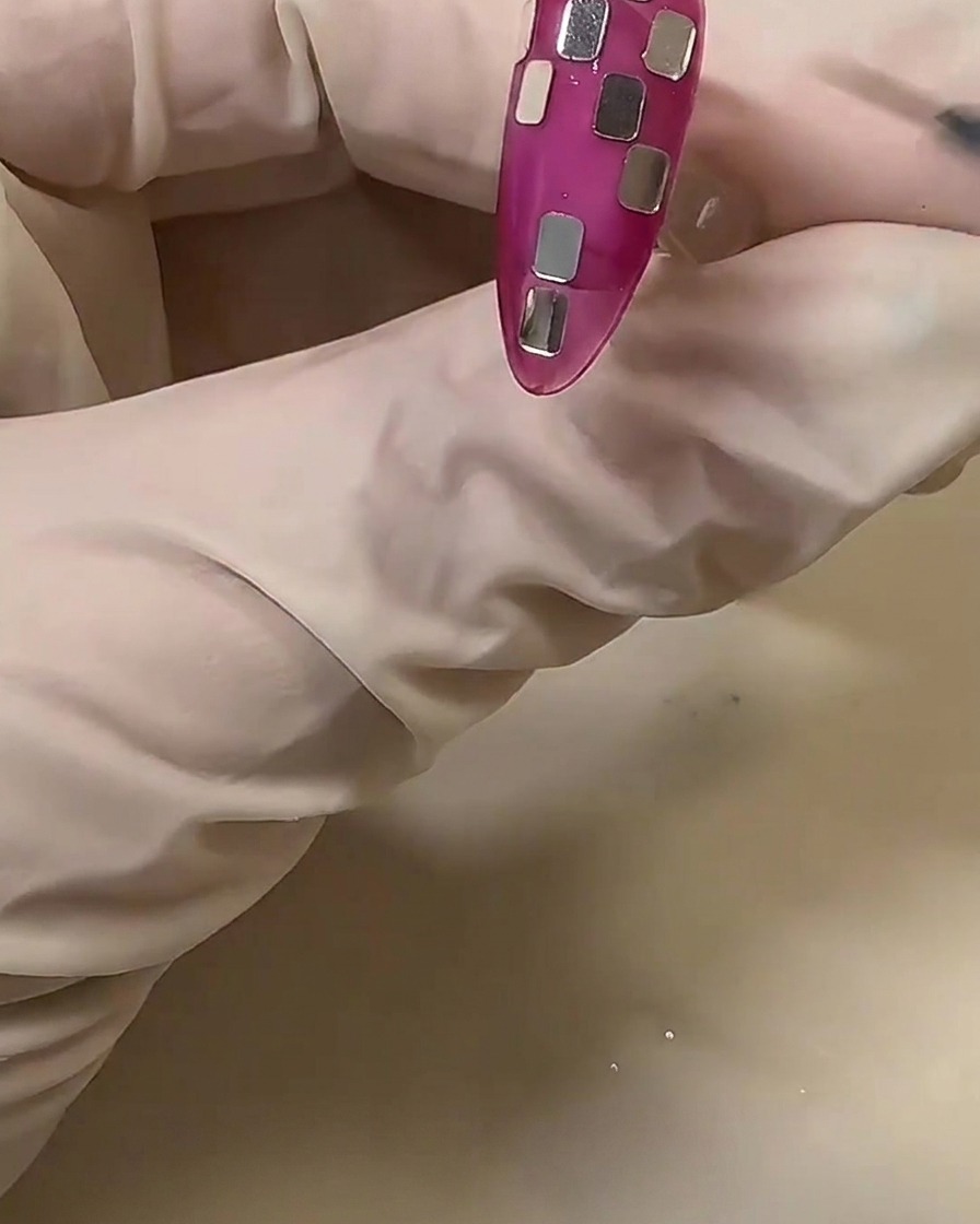 Une main gantée ajustant de petits morceaux carrés de feuille argentée sur un ongle rose translucide pour créer un motif en damier.