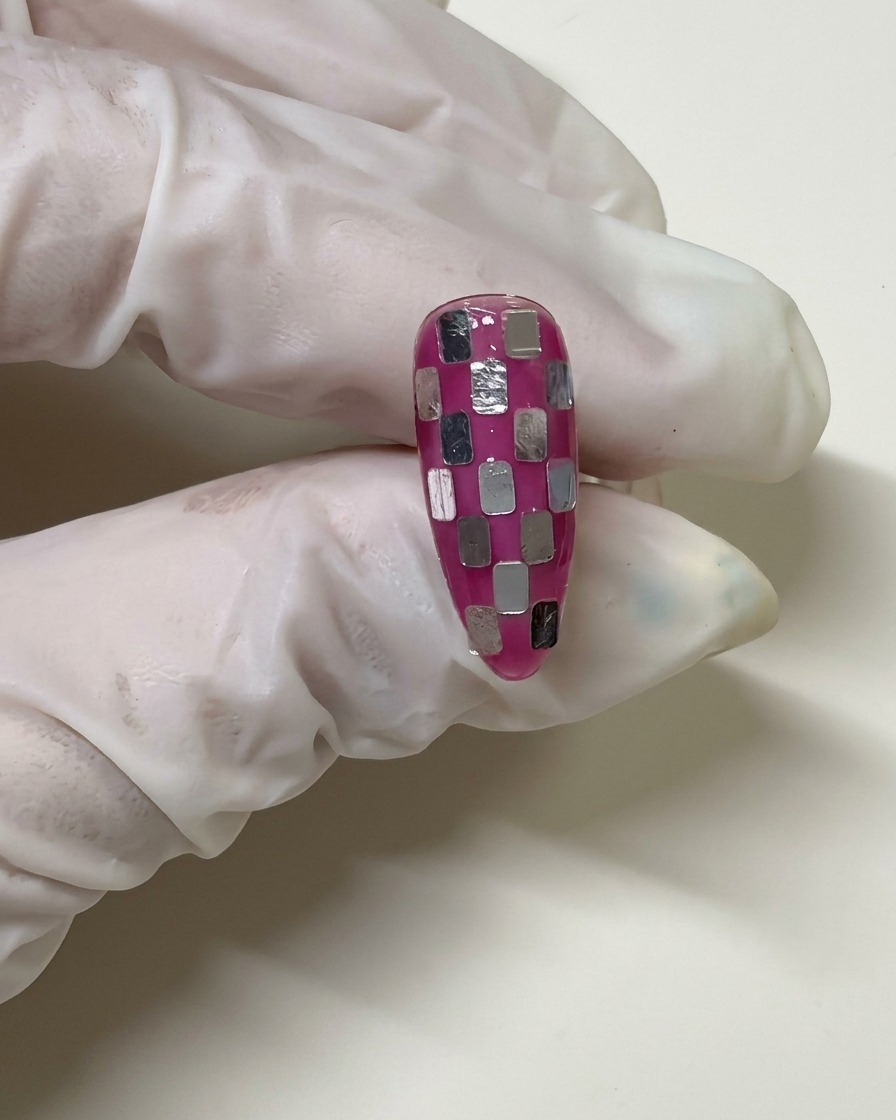 Une main gantée tenant un ongle avec un motif en damier qui a été recouvert dune fine couche de gel rose pour fondre les carrés argentés.