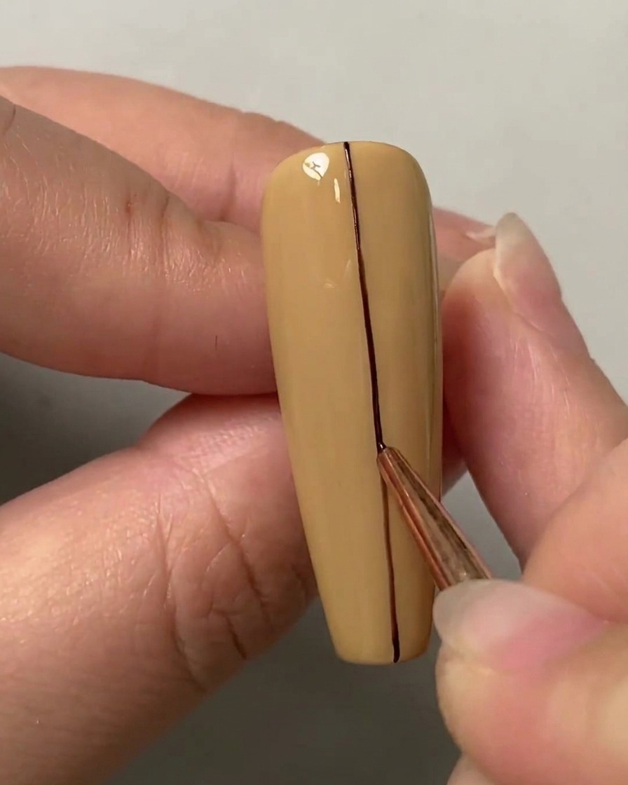 Un pinceau fin traçant une ligne droite sombre au centre vertical dun ongle de couleur nude.