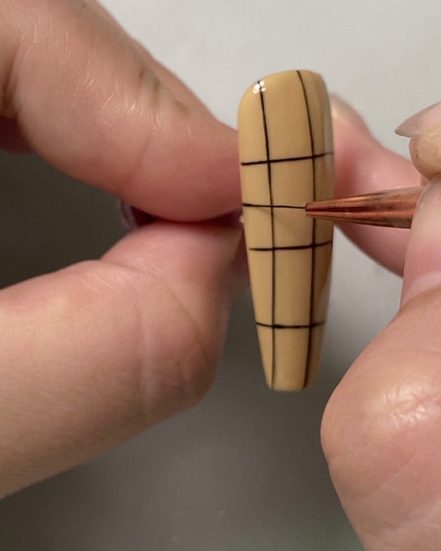 Un pinceau fin ajoutant une ligne horizontale supplémentaire à la partie supérieure dun motif quadrillé sur un ongle.