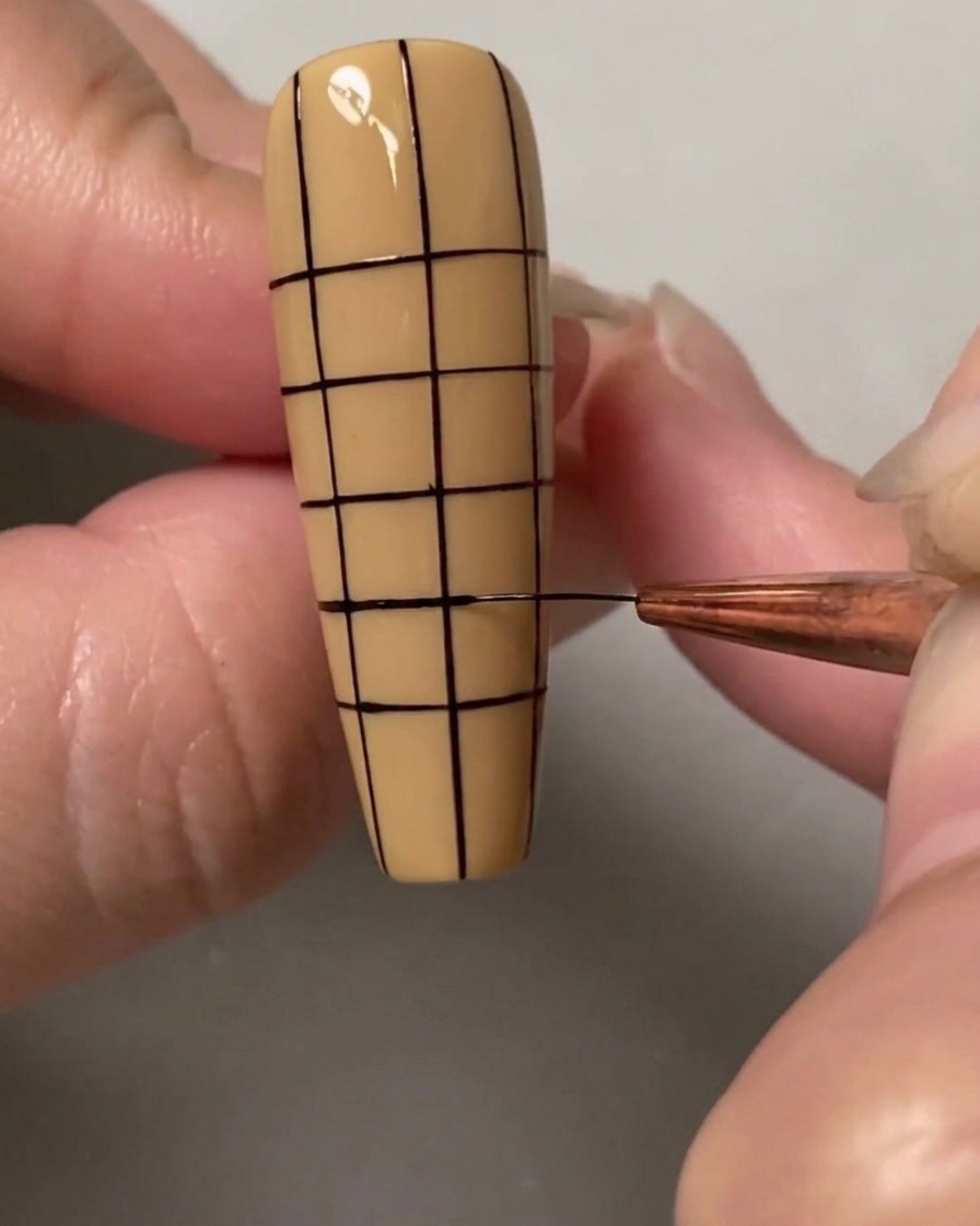 Un pinceau de nail art traçant les dernières lignes horizontales pour former un quadrillage petit et régulier sur tout longle.