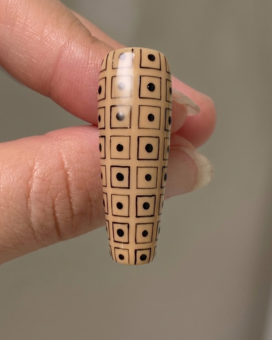 Un ongle long de couleur nude avec un quadrillage noir dessiné à la main, avec des points noirs alternés à lintérieur des carrés.