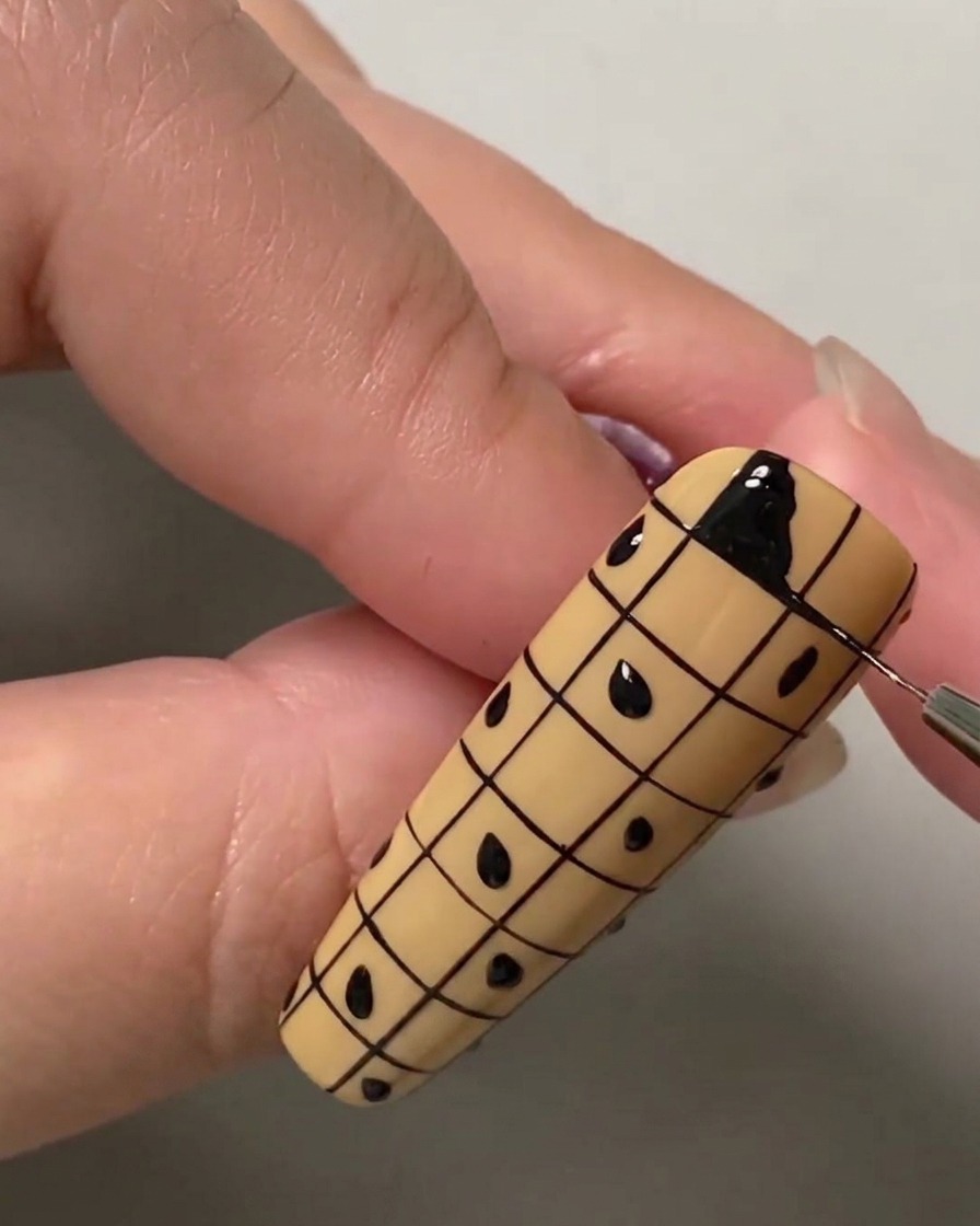 Gros plan dun pinceau fin remplissant le carré supérieur gauche dun quadrillage sur un ongle nude avec du vernis gel noir.