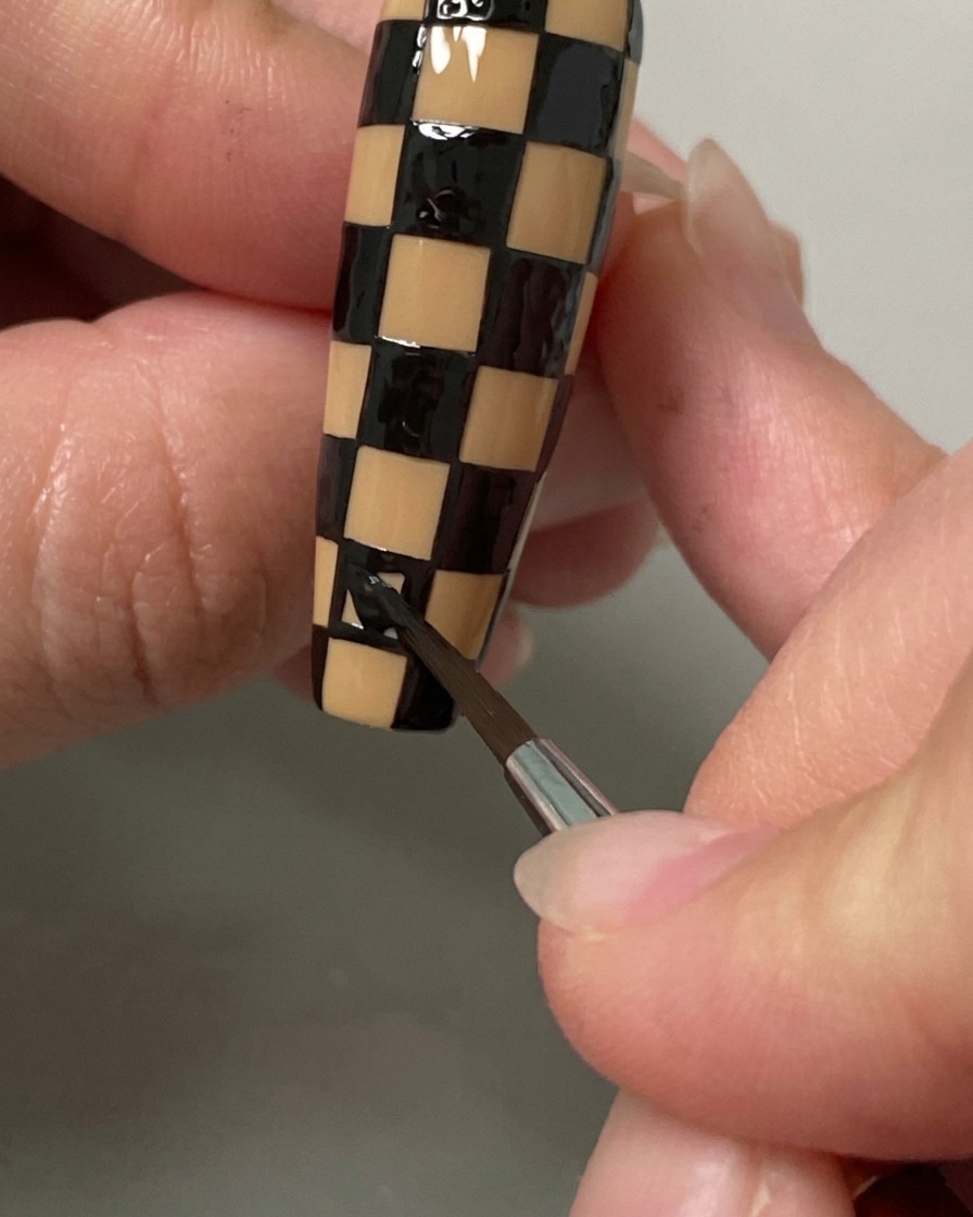 Un pinceau fin remplissant les derniers carrés dun motif damier noir et nude sur un ongle long.