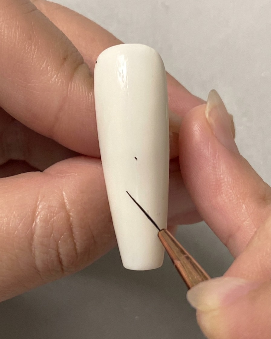 Une main utilisant un pinceau fin pour placer un petit point sur un ongle blanc uni.