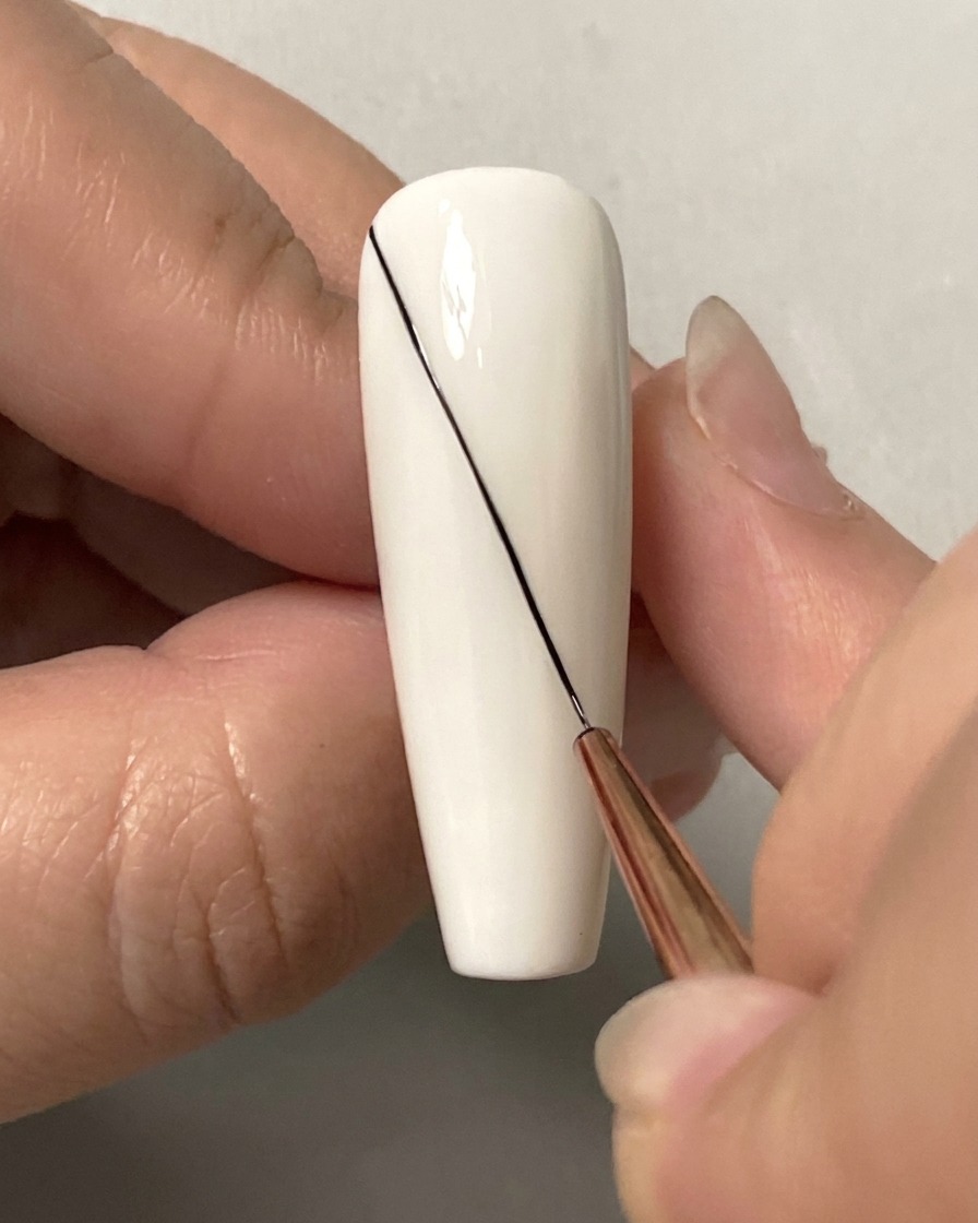 Gros plan dun pinceau fin dessinant une longue ligne noire diagonale sur un ongle blanc.