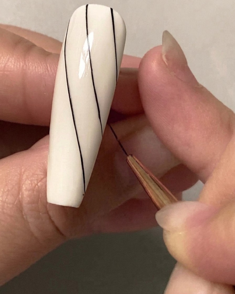 Gros plan dun pinceau fin de nail art dessinant une deuxième ligne noire diagonale parallèle sur la moitié supérieure dun ongle blanc.