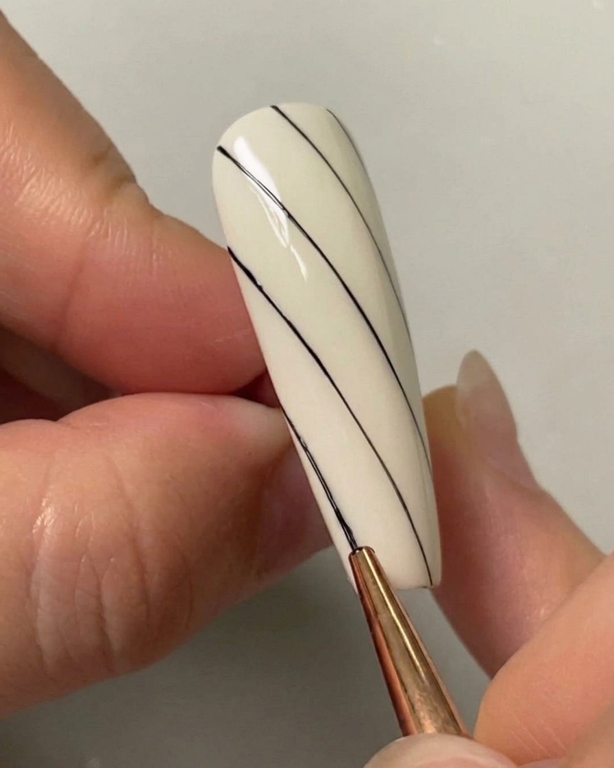 Un pinceau de nail art dessinant une troisième ligne noire diagonale sur le côté inférieur gauche dun ongle peint en blanc.