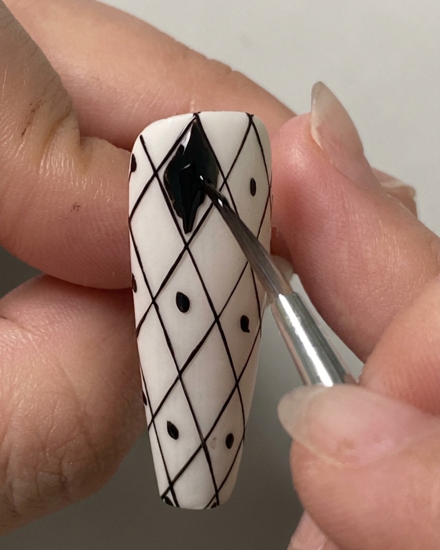 Un pinceau fin remplissant soigneusement la forme en losange la plus haute avec du vernis gel noir sur un quadrillage blanc pré-dessiné.