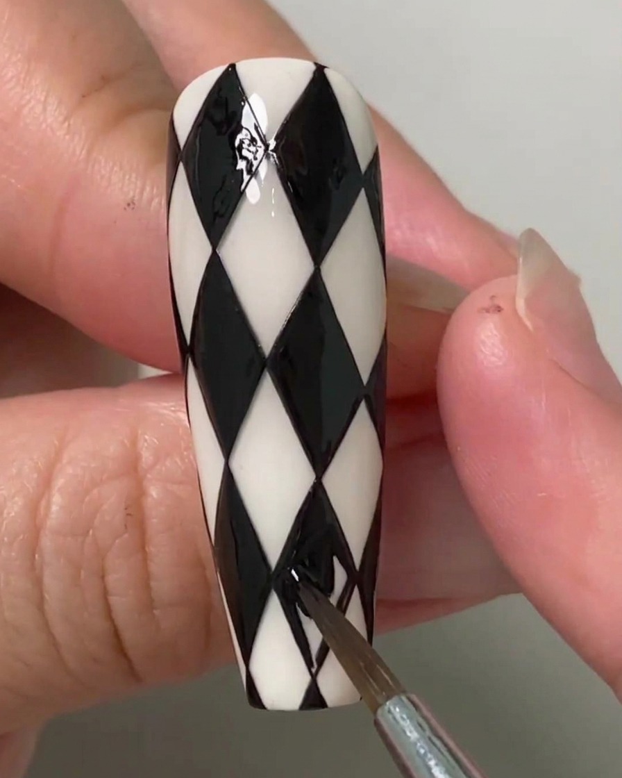 Un artiste ongulaire utilisant un pinceau fin pour remplir les dernières formes en losange sur un ongle à motif argyle noir et blanc.