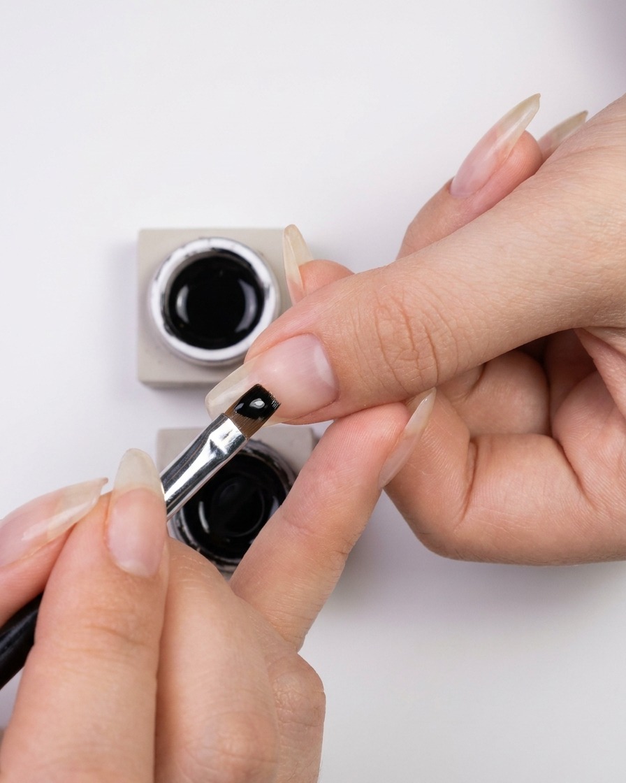 Une main trempant un pinceau liner fin pour nail art dans un petit pot de vernis gel noir brillant.