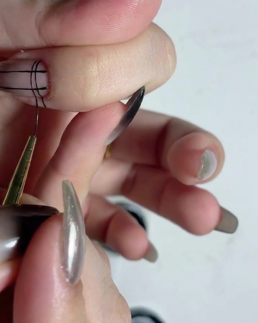 Un pinceau liner fin traçant des lignes horizontales noires sur des lignes verticales pour créer une grille sur un ongle.