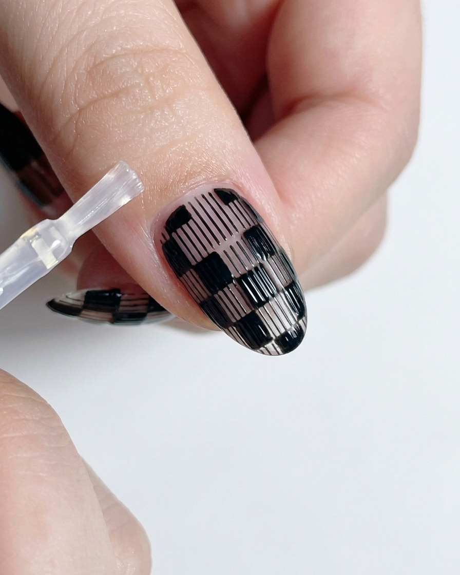 Un pinceau à top coat appliquant du vernis brillant transparent sur un ongle à damier noir et blanc terminé.