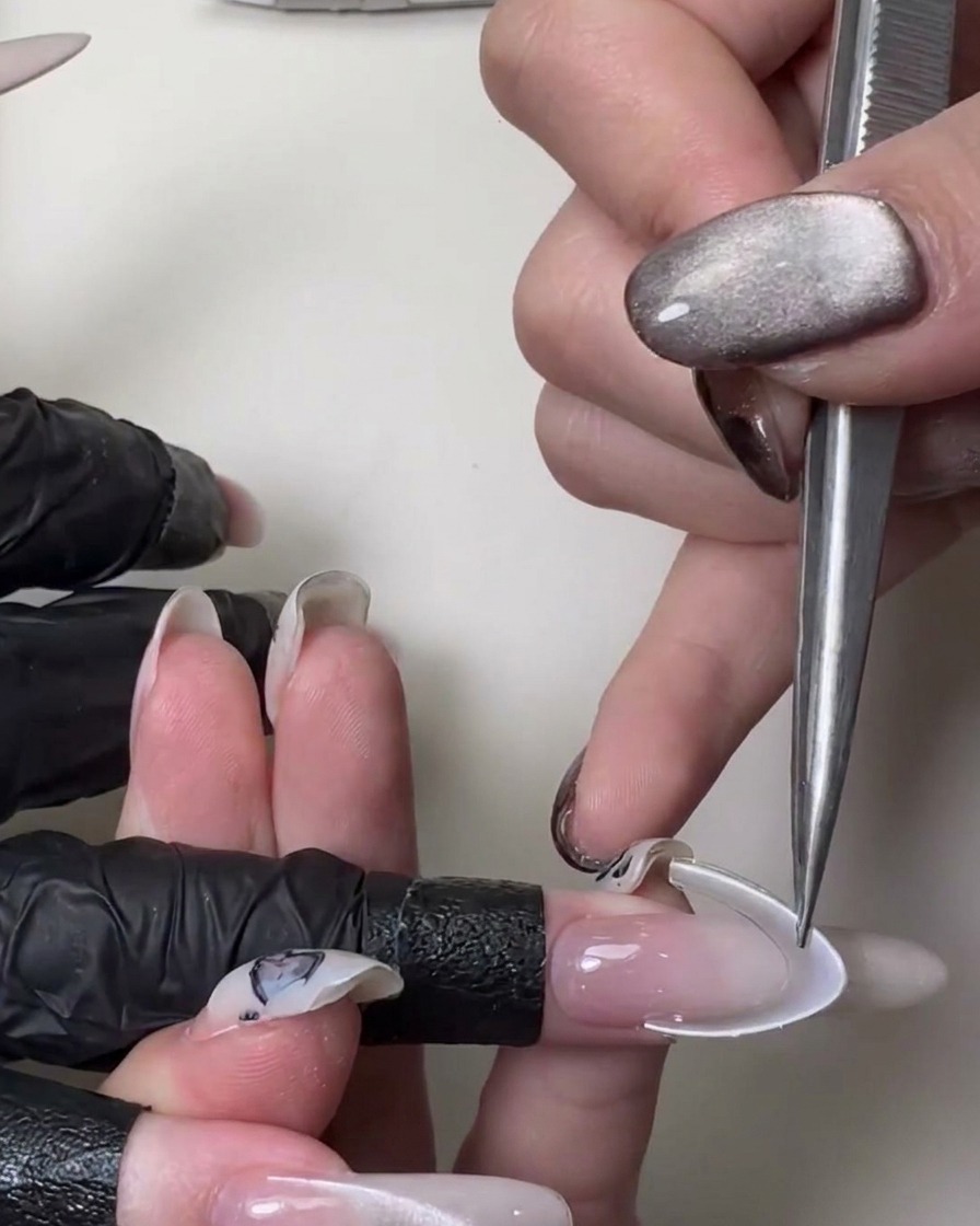 Gros plan dune main utilisant des pincettes en métal pour appliquer un autocollant blanc courbé pour ligne de sourire French sur un long ongle stiletto rose translucide.