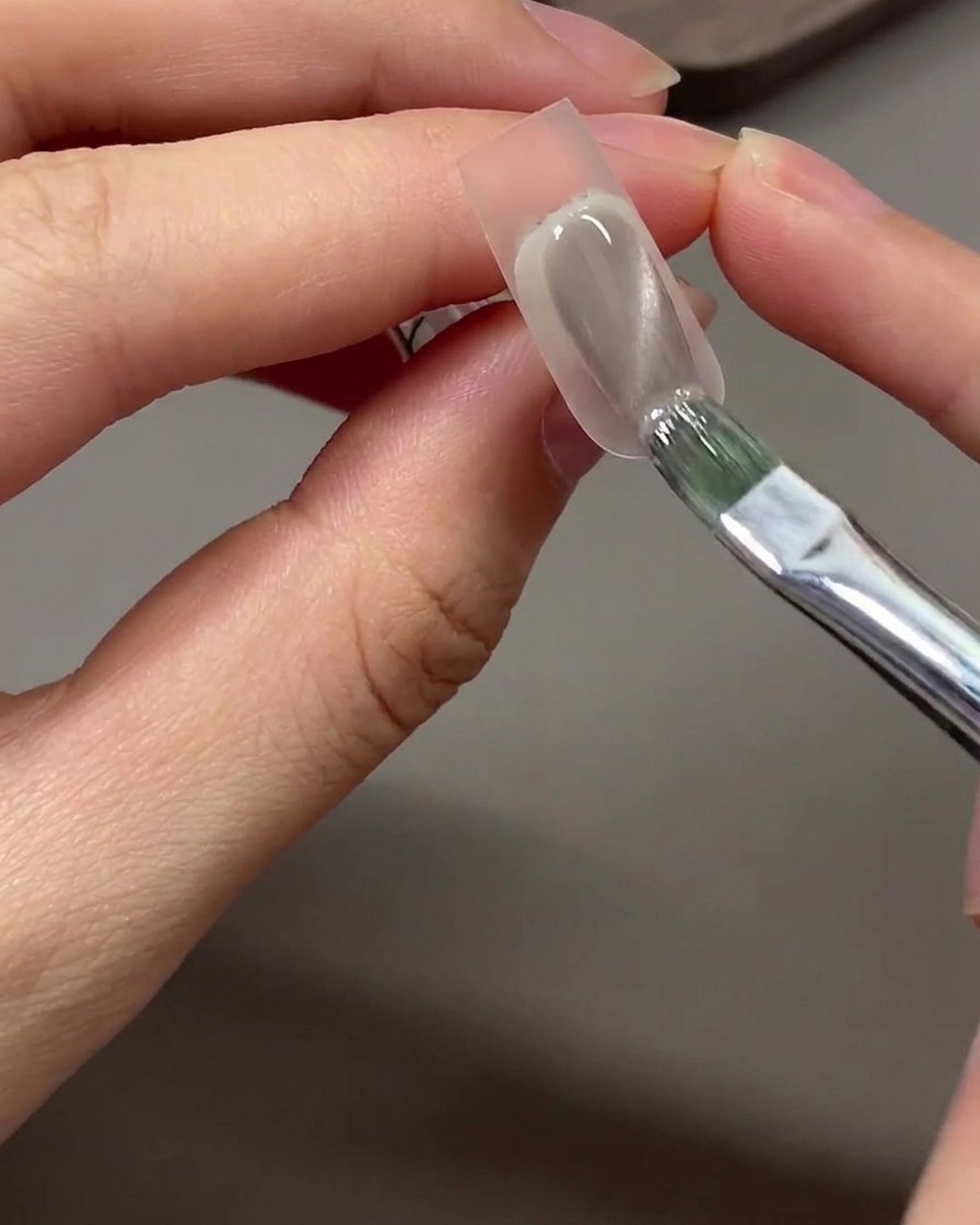 Un pinceau appliquant du gel cat eye gris-nude sur la zone en dégradé tapotée à léponge sur un ongle.