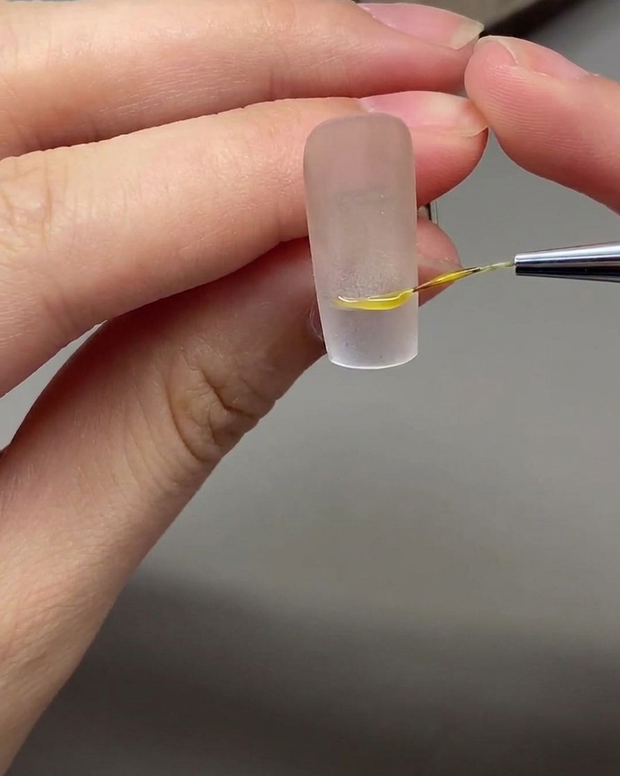 Un pinceau de nail art fin dessinant une ligne de sourire French blanche et précise sur un ongle à base nude.