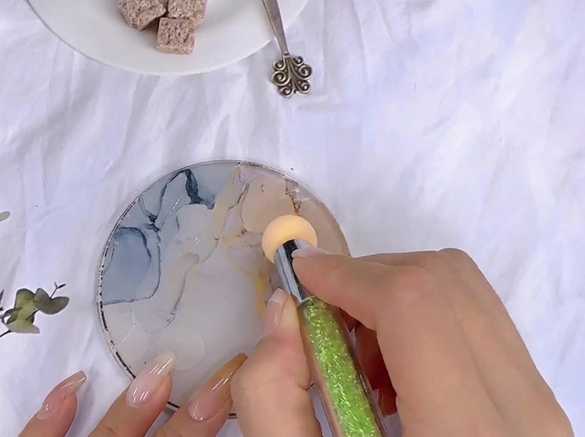 Une éponge de maquillage tapotant dans du gel de couleur étalé sur une palette en verre à motifs.