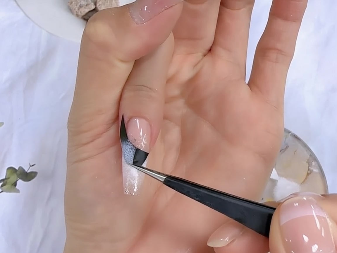 Une paire de pinces retirant délicatement un sticker guide français noir dun ongle pour révéler le motif dégradé en dessous.